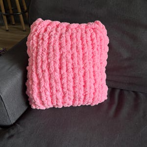 Peut inclure: Un coussin carr&eacute; rose en tricot &eacute;pais repose sur un canap&eacute; gris fonc&eacute;. Le coussin est fait d'un fil doux et textur&eacute;, et sa couleur vive contraste avec le fond plus sombre. Les dimensions du coussin sont d'environ 35 cm sur 35 cm.