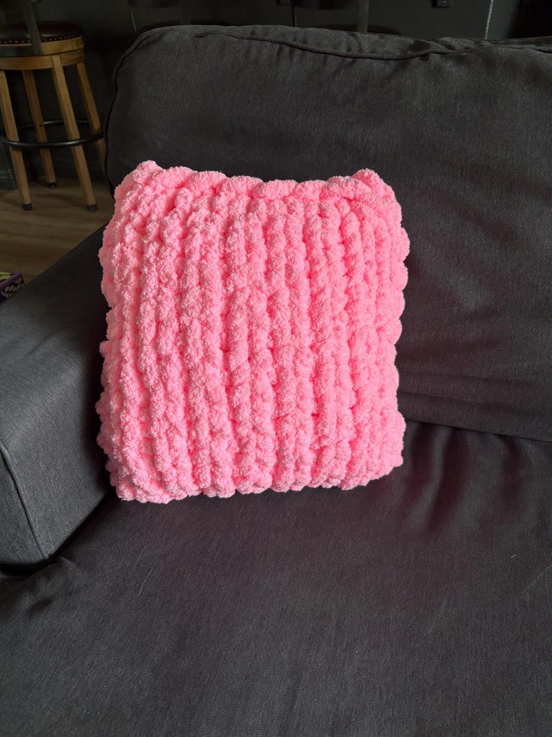 Peut inclure: Un coussin carr&eacute; rose en tricot &eacute;pais repose sur un canap&eacute; gris fonc&eacute;. Le coussin est fait d'un fil doux et textur&eacute;, et sa couleur vive contraste avec le fond plus sombre. Les dimensions du coussin sont d'environ 35 cm sur 35 cm.