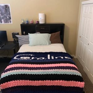 K&ouml;nnte beinhalten: Eine handgestrickte Decke mit horizontalen Streifen in Marineblau, Rosa, Schwarz und Hellblau, mit dem Wort "Calico" in Wei&szlig; eingestrickt. Die Decke liegt auf einem Bett mit einem schwarzen Kopfteil. Der Raum hat eine neutrale Farbgebung.