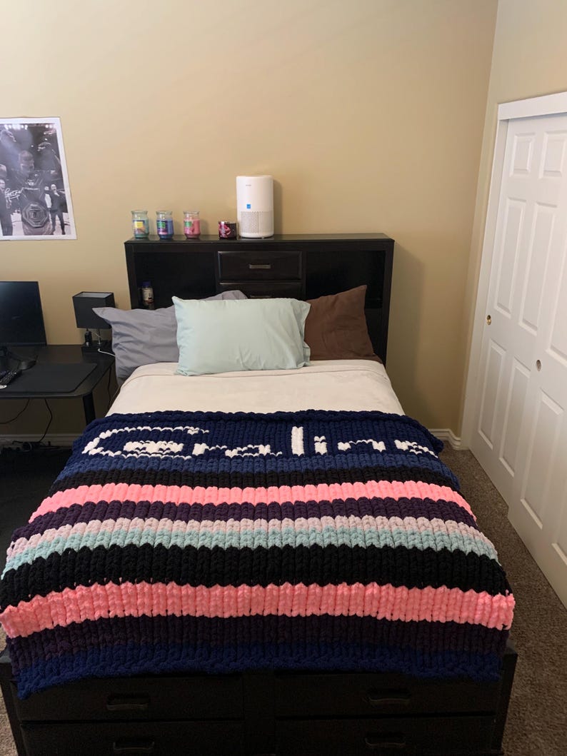 K&ouml;nnte beinhalten: Eine handgestrickte Decke mit horizontalen Streifen in Marineblau, Rosa, Schwarz und Hellblau, mit dem Wort "Calico" in Wei&szlig; eingestrickt. Die Decke liegt auf einem Bett mit einem schwarzen Kopfteil. Der Raum hat eine neutrale Farbgebung.