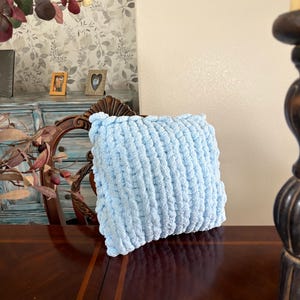 Peut inclure: Un coussin en tricot &eacute;pais bleu clair. Le coussin est de forme carr&eacute;e et pr&eacute;sente une surface textur&eacute;e et boucl&eacute;e. Le coussin repose sur une surface en bois fonc&eacute;.