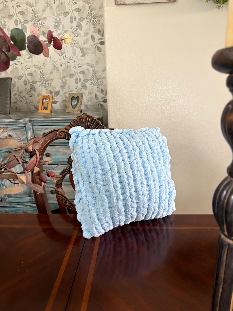 Peut inclure: Un coussin en tricot &eacute;pais bleu clair. Le coussin est de forme carr&eacute;e et pr&eacute;sente une surface textur&eacute;e et boucl&eacute;e. Le coussin repose sur une surface en bois fonc&eacute;.