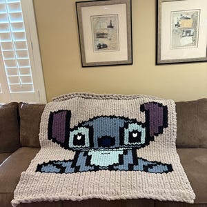 Puede incluir: Una manta de punto grueso con una imagen pixelada de Stitch de Lilo & Stitch. La manta es principalmente gris claro con detalles en azul, morado y negro. La manta est&aacute; sobre un sof&aacute; marr&oacute;n.