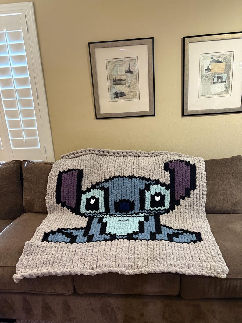 Puede incluir: Una manta de punto grueso con una imagen pixelada de Stitch de Lilo & Stitch. La manta es principalmente gris claro con detalles en azul, morado y negro. La manta est&aacute; sobre un sof&aacute; marr&oacute;n.