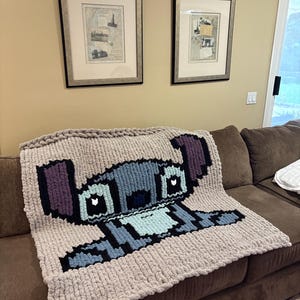 Puede incluir: Una manta de punto grueso con un dise&ntilde;o pixelado del personaje Stitch de Lilo & Stitch. La manta es principalmente beige con detalles azules, negros y morados. Se encuentra sobre un sof&aacute; marr&oacute;n.