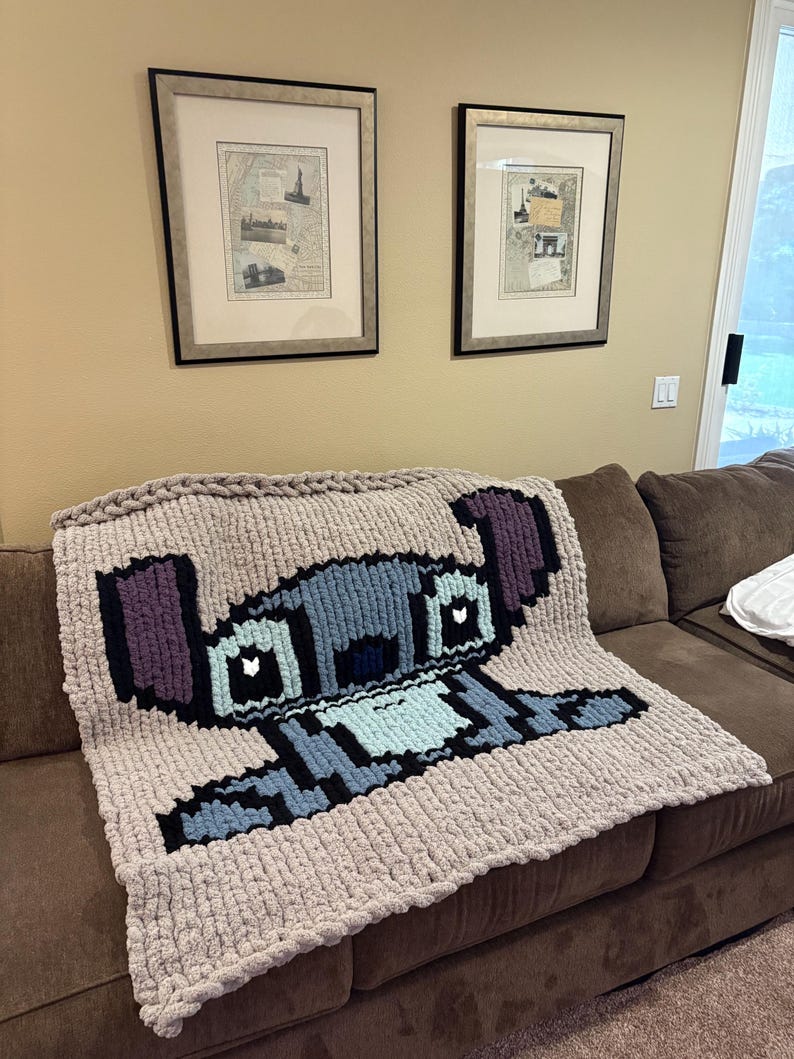 Puede incluir: Una manta de punto grueso con un dise&ntilde;o pixelado del personaje Stitch de Lilo & Stitch. La manta es principalmente beige con detalles azules, negros y morados. Se encuentra sobre un sof&aacute; marr&oacute;n.