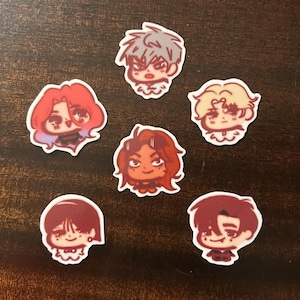May include: A collection of six anime-style stickers featuring stylized portraits of characters with various hair colors and expressions. The stickers have a white border and are arranged on a dark brown surface.