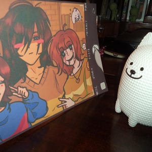 May include: An illustrated print featuring three anime-style characters with red hair and various expressions. A white, textured dog-like figurine with a smiling face is to the right of the print. The print has text on the side.