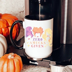 May include: White coffee mug with a black handle and a black rim. The mug has a graphic of three witches with the text "ZERO AMUCKS GIVEN".