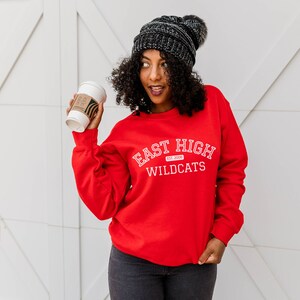 May include: A woman wearing a red sweatshirt with the text "East High Est. 2008 Wildcats" in white. She is holding a coffee cup in her right hand and wearing a black beanie with a pom pom.