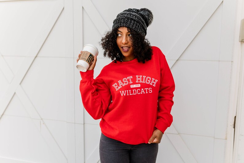 May include: A woman wearing a red sweatshirt with the text "East High Est. 2008 Wildcats" in white. She is holding a coffee cup in her right hand and wearing a black beanie with a pom pom.