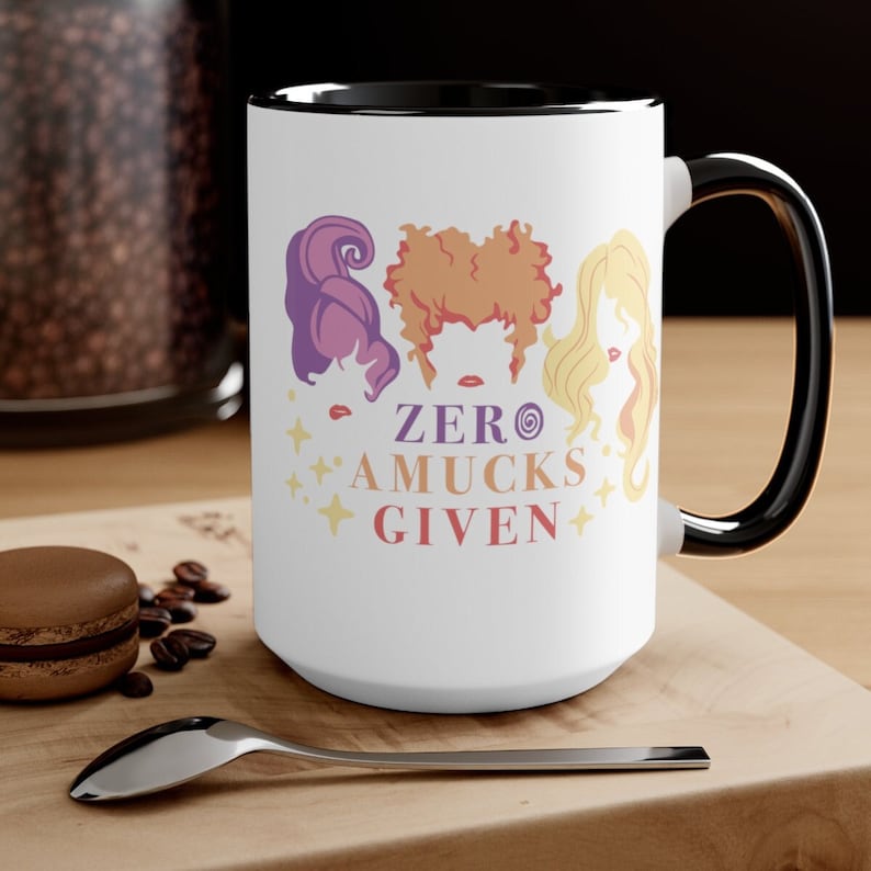May include: White ceramic mug with a black rim and handle. The mug features a graphic of three witches with the text "ZERO AMUCKS GIVEN".