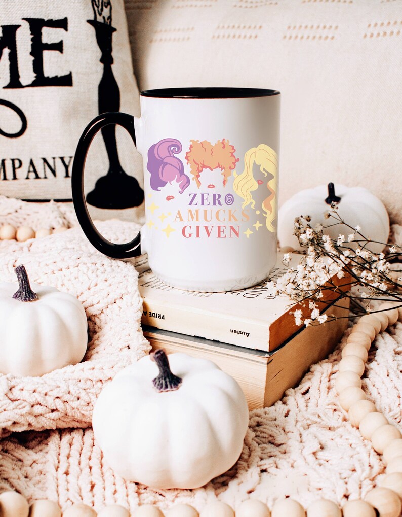 May include: White ceramic mug with a black handle and black rim. The mug has a graphic of three women with the text "Zero A-Mucks Given". The mug is sitting on a white knitted blanket with white pumpkins and a book.