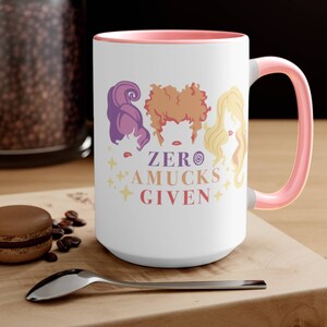 May include: White ceramic coffee mug with a pink rim and handle. The mug features a graphic of three witches with the text "ZERO AMUCKS GIVEN".