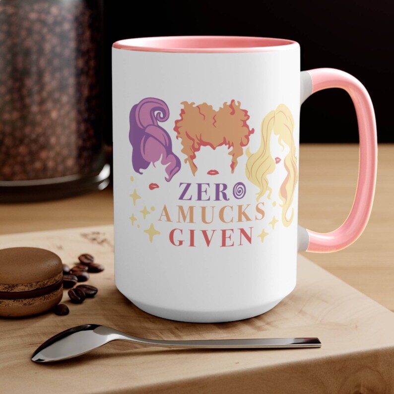 May include: White ceramic coffee mug with a pink rim and handle. The mug features a graphic of three witches with the text "ZERO AMUCKS GIVEN".