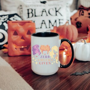 May include: White ceramic coffee mug with a black rim and handle. The mug features a colorful illustration of three witches with the text "Zero Mucks Given".