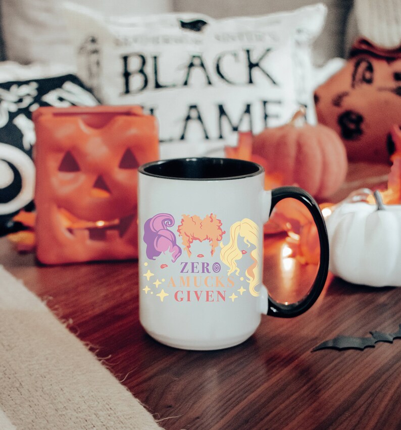 May include: White ceramic coffee mug with a black rim and handle. The mug features a colorful illustration of three witches with the text "Zero Mucks Given".