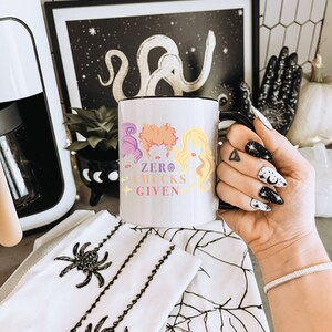 May include: White ceramic mug with a black handle. The mug features a colorful illustration of three witches with the text "ZERO MUCKS GIVEN".