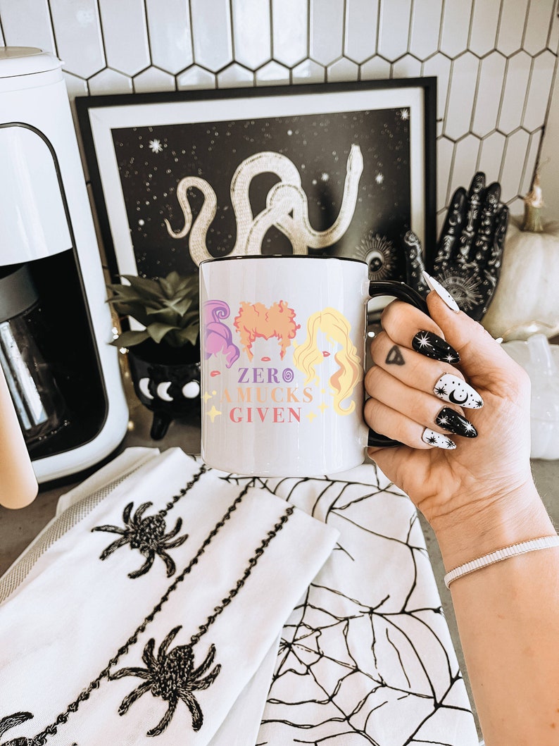 May include: White ceramic mug with a black handle. The mug features a colorful illustration of three witches with the text "ZERO MUCKS GIVEN".