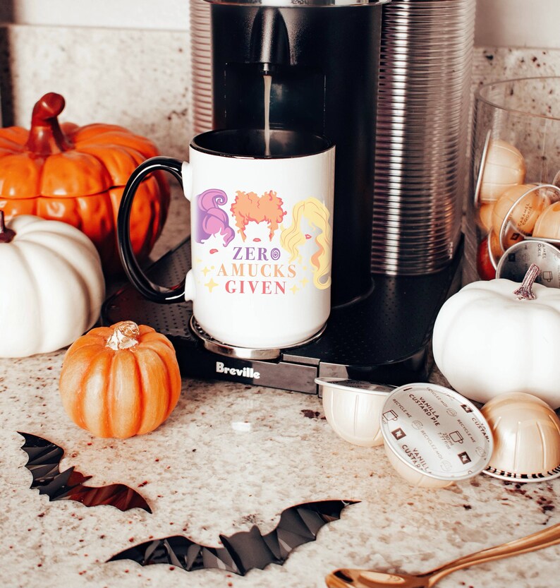 May include: White coffee mug with a black handle and a black rim. The mug has a graphic of three witches with the text "ZERO AMUCKS GIVEN".
