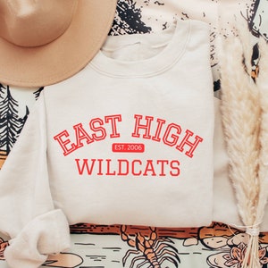 May include: A light beige sweatshirt with the text "East High" in red with a red banner underneath that says "Est. 2006" and "Wildcats" in red below the banner.