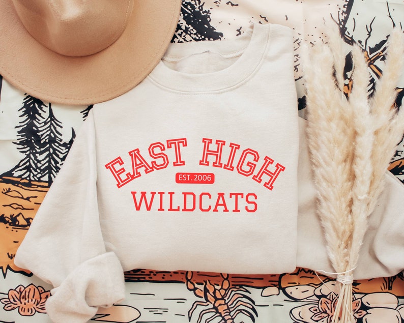 May include: A light beige sweatshirt with the text "East High" in red with a red banner underneath that says "Est. 2006" and "Wildcats" in red below the banner.