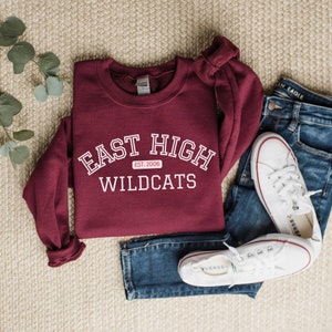 May include: A maroon sweatshirt with white text that reads "East High Wildcats Est. 2006". The sweatshirt is laid out on a beige carpet with a pair of blue jeans and white trainers.