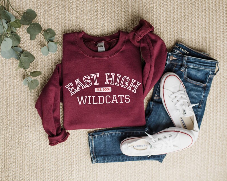 May include: A maroon sweatshirt with white text that reads "East High Wildcats Est. 2006". The sweatshirt is laid out on a beige carpet with a pair of blue jeans and white trainers.
