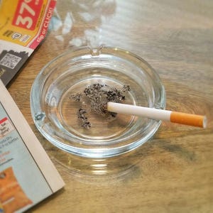 May include: A clear glass ashtray with a cigarette butt and ash inside. The ashtray is sitting on a wooden surface.