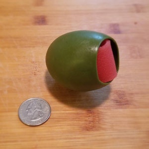 May include: A green rubber dog toy shaped like an olive with a red rubber insert. The toy is about the size of a quarter.