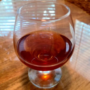 May include: A clear glass with a round base filled with a dark red liquid. The glass is sitting on a wooden surface.