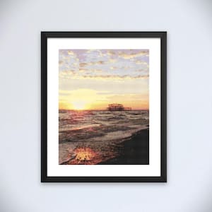 Könnte beinhalten: Gerahmter Druck eines Sonnenuntergangs über dem Meer. Das Bild zeigt eine leuchtend gelbe Sonne, die über dem Wasser untergeht, mit einem Pier in der Ferne. Der Himmel ist eine Mischung aus Gelb, Orange und Lila. Der Druck ist in einem schwarzen Rahmen.