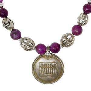 May include: A necklace featuring large, deep purple beads interspersed with silver filigree beads and smaller purple beads. A silver pendant with a detailed design hangs at the center. The necklace is on a white background.