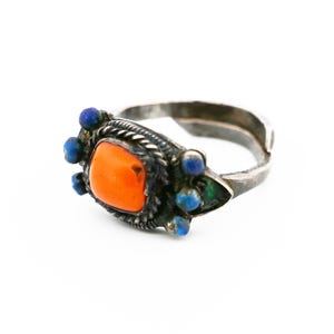 Puede incluir: Un anillo de plata con una piedra naranja engastada en un engaste de plata. El anillo está decorado con pequeñas piedras azules.