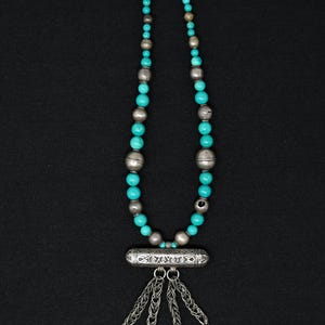 May include: A silver necklace with turquoise and silver beads. The necklace has a cylindrical pendant with etched designs and dangling chains with square charms. The necklace is displayed against a black background.