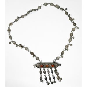 May include: A silver-toned necklace with a central rectangular pendant featuring three oval, reddish-orange stones. The necklace has a delicate chain with floral and teardrop-shaped links, and small dangling charms.