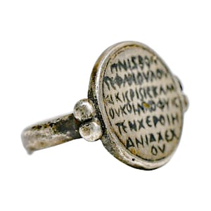 May include: A silver-toned metal ring featuring a large, oval-shaped face with engraved dark lettering. The band connects to the oval face with two small, rounded decorative spheres on each side. The surface shows signs of age and wear.