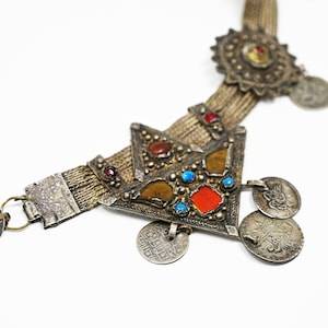 May include: A silver chain necklace with a triangular pendant decorated with red, orange, and blue stones. The pendant is surrounded by several silver coins with Arabic script.