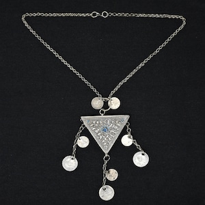 May include: Silver-toned necklace with a triangular pendant and dangling coin charms. The pendant features intricate detailing and a central blue accent. The necklace has a delicate chain and is set against a black background.