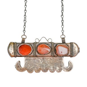 May include: A silver-toned metal pendant necklace featuring a cylindrical bar set with three oval-shaped orange stones. The pendant includes a decorative, curved metal plate with intricate embossed patterns hanging from the base and two chain links.