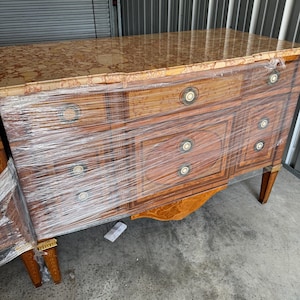 Puede incluir: Un antiguo cofre de cajones de madera con tapa de mármol. Los cajones están hechos de madera con un diseño de marquetería y tienen tiradores de latón. El cofre está envuelto en plástico.