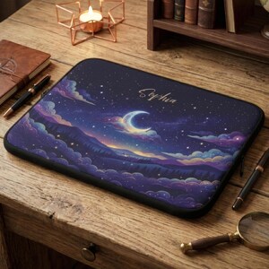 May include: A laptop sleeve featuring a celestial design with a crescent moon, stars, and clouds in shades of purple, blue, and orange. The name "Sophia" is written in gold script. The sleeve is on a wooden surface.