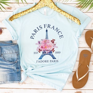 May include: Light blue t-shirt with a graphic of the Eiffel Tower surrounded by pink and purple flowers. The text "Paris France" arches above the tower, with "Est 1889" and "J'adore Paris" below. The shirt is displayed on a gold hanger.