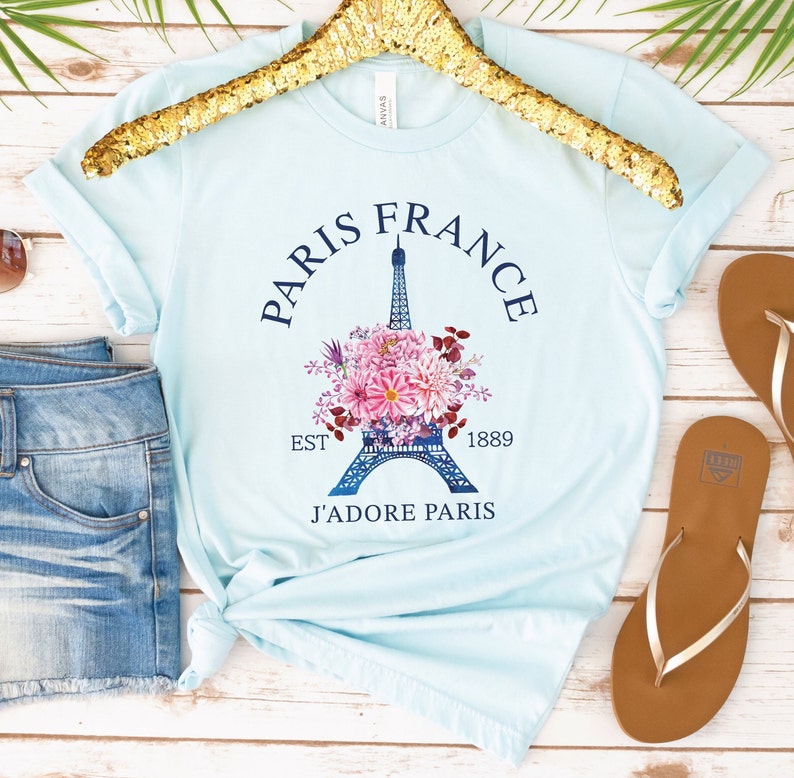 May include: Light blue t-shirt with a graphic of the Eiffel Tower surrounded by pink and purple flowers. The text "Paris France" arches above the tower, with "Est 1889" and "J'adore Paris" below. The shirt is displayed on a gold hanger.