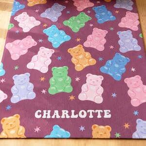 Puede incluir: Una alfombra rectangular con fondo morado oscuro que presenta un patrón repetitivo de ositos de goma coloridos en tonos rosa, azul, verde y naranja, intercalados con pequeñas estrellas. El nombre CHARLOTTE está impreso en letras blancas.