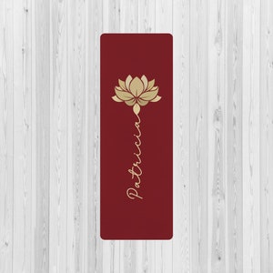 May include: A rectangular yoga mat in a deep burgundy red color. The mat features a gold-colored graphic of a lotus flower at the top, with the name Patricia written in a gold-colored cursive script extending vertically downward from the flower.