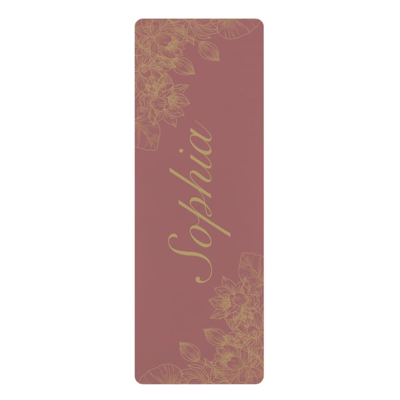 May include: A rectangular yoga mat in a dusty rose color featuring a gold-toned line art floral pattern in the top left and bottom right corners. The name Sophia is printed in a large, gold-toned cursive script font vertically along the center.