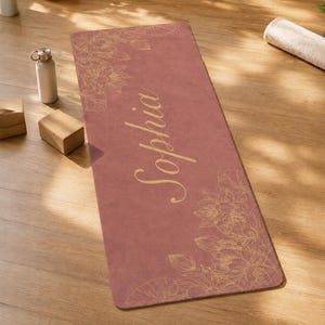 May include: A rectangular yoga mat in a dusty rose color featuring a gold-toned line art floral pattern in the top left and bottom right corners. The name Sophia is printed in a large, gold-toned cursive script across the center of the mat.