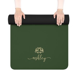 May include: A dark green yoga mat featuring a gold lotus flower icon centered above the name ashley written in a cursive script font. The top portion of the mat is rolled into a black cylinder, displaying a smooth, solid-colored surface.