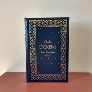 May include: A hardcover book titled "Charles Dickens Four Complete Novels." The book has a dark blue cover with gold decorative borders and text. The book is standing upright on a red surface.